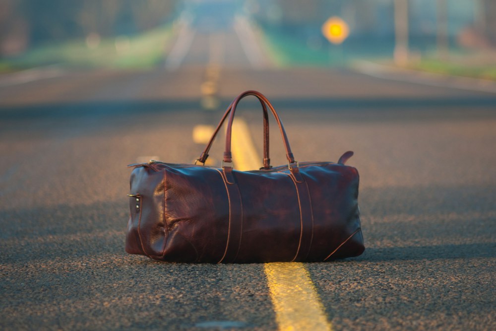 brown leather duffel bag in middle on gray asphalt road; illustrative stock photograph