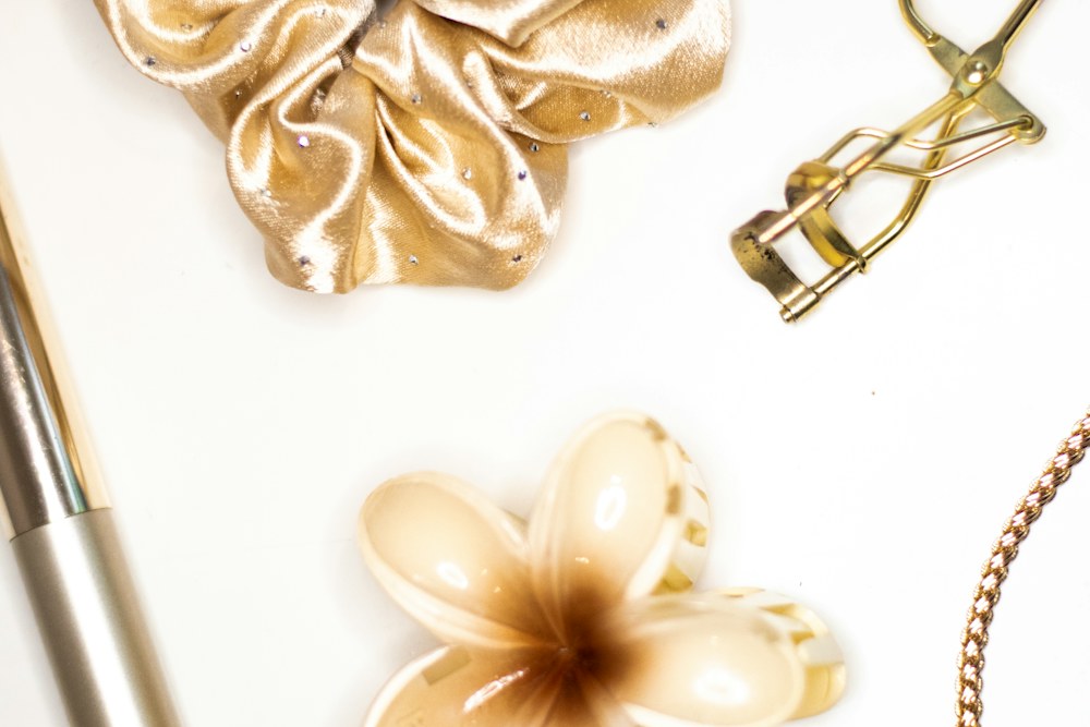 A gold scrunchie, eyelash curler, and flower hair clip on a white surface; illustrative stock photograph