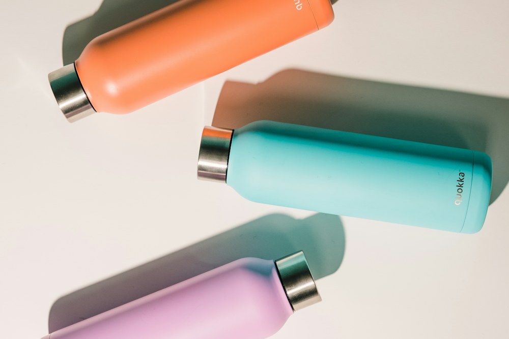 Three colorful metal water bottles on a white surface with long shadows; illustrative stock photograph